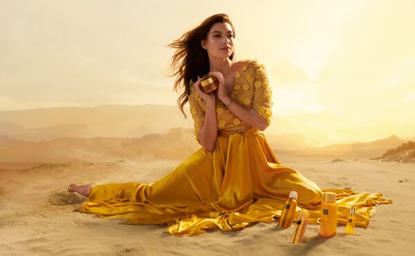 Woman in a flowing yellow dress holding The Ritual of Mehr skincare products, sitting on sand against a golden sky backdrop.